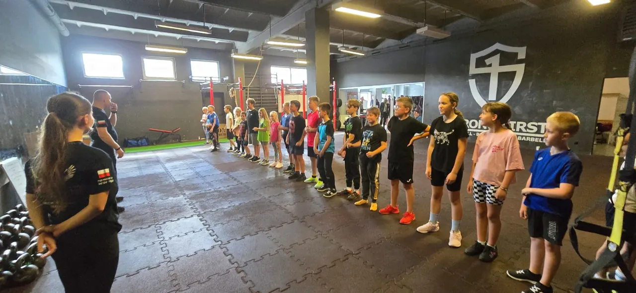 Dzieci w szeregu na sali treningowej w obecności instruktorów, obok odważników kettlebell
